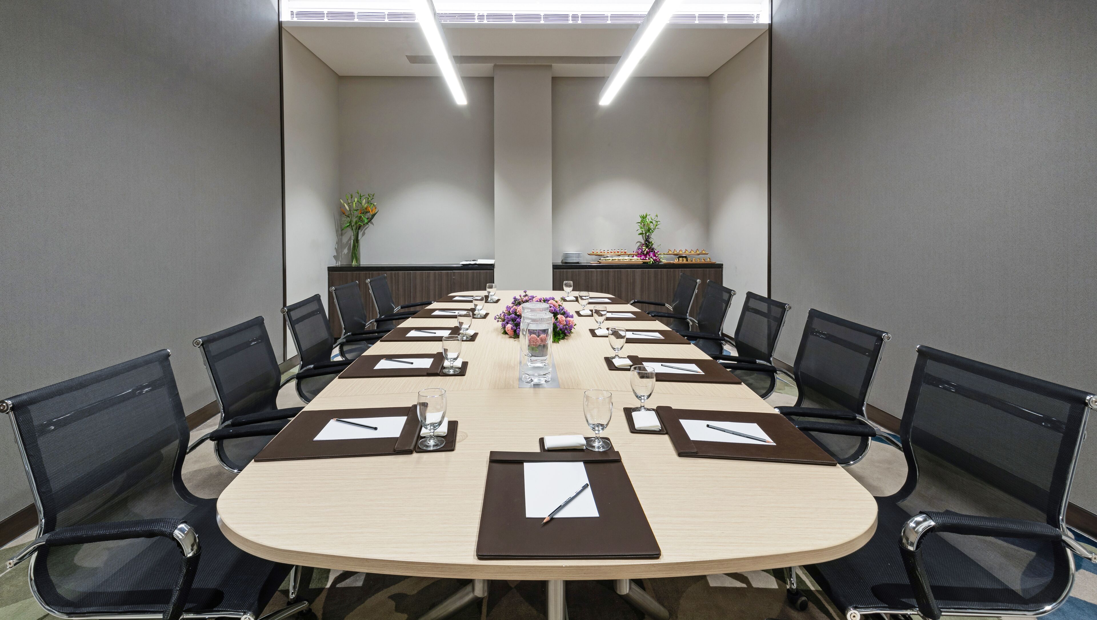 Een vergaderzaal met een lange tafel, zwarte mesh-stoelen en een bloemenarrangement.