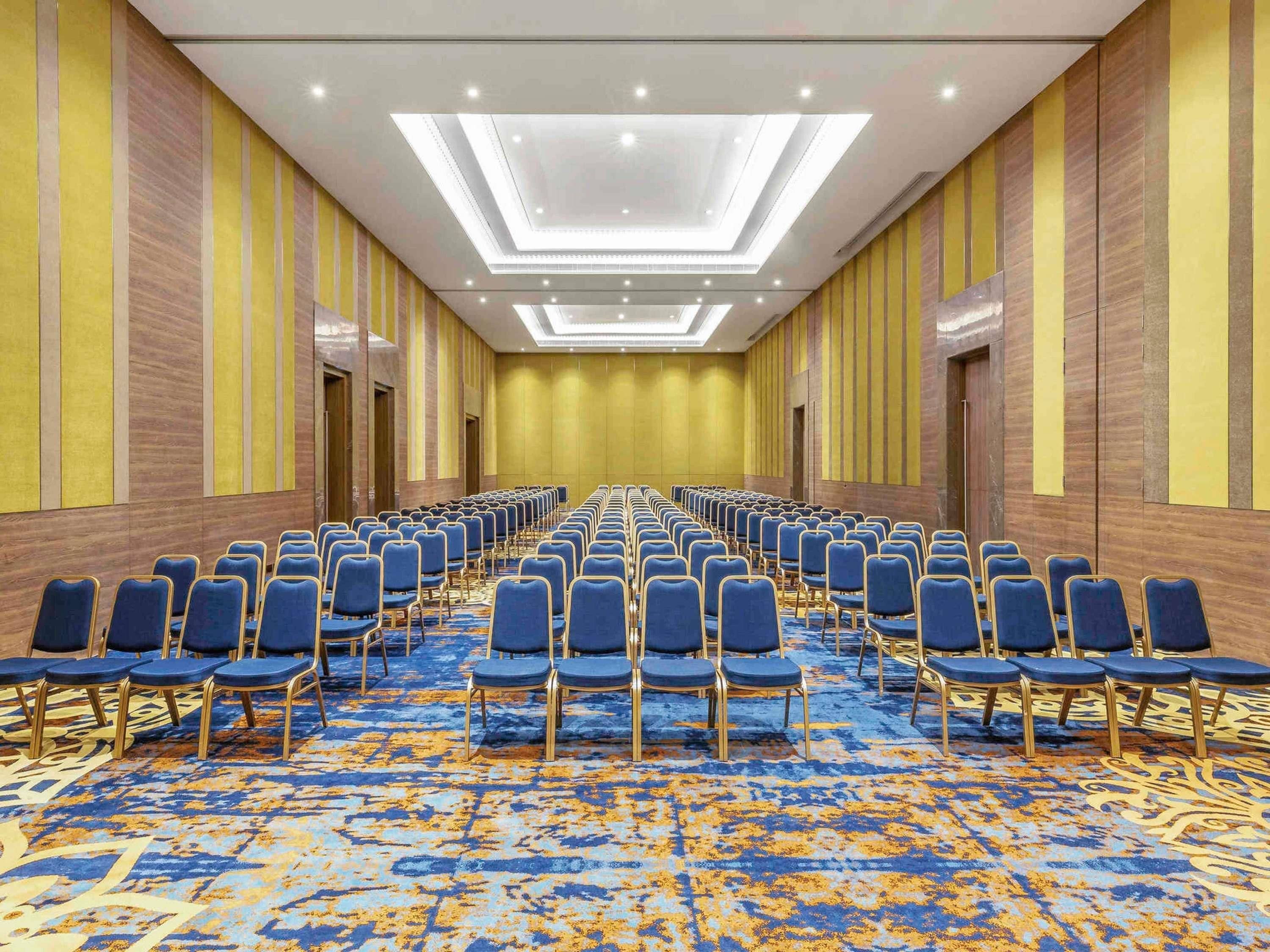 Un salón de conferencias con filas de sillas azules, alfombra estampada y un diseño moderno en el techo.