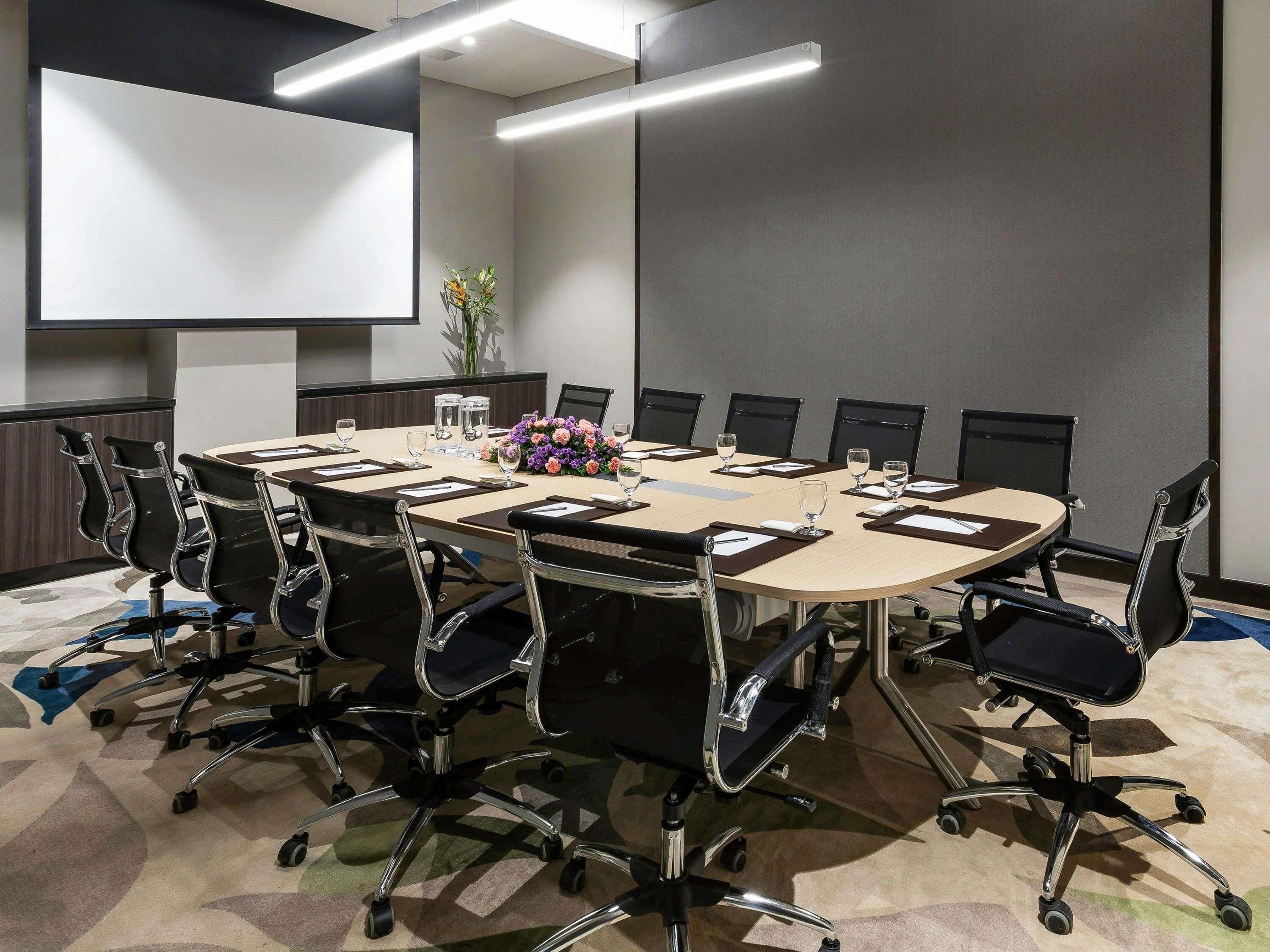 A conference room with a large oval table, black chairs, a projector screen, and a floral centerpiece.