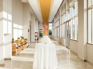 Un pasillo de hotel amplio con una larga mesa y sillas, una exhibición de alimentos y ventanas grandes.