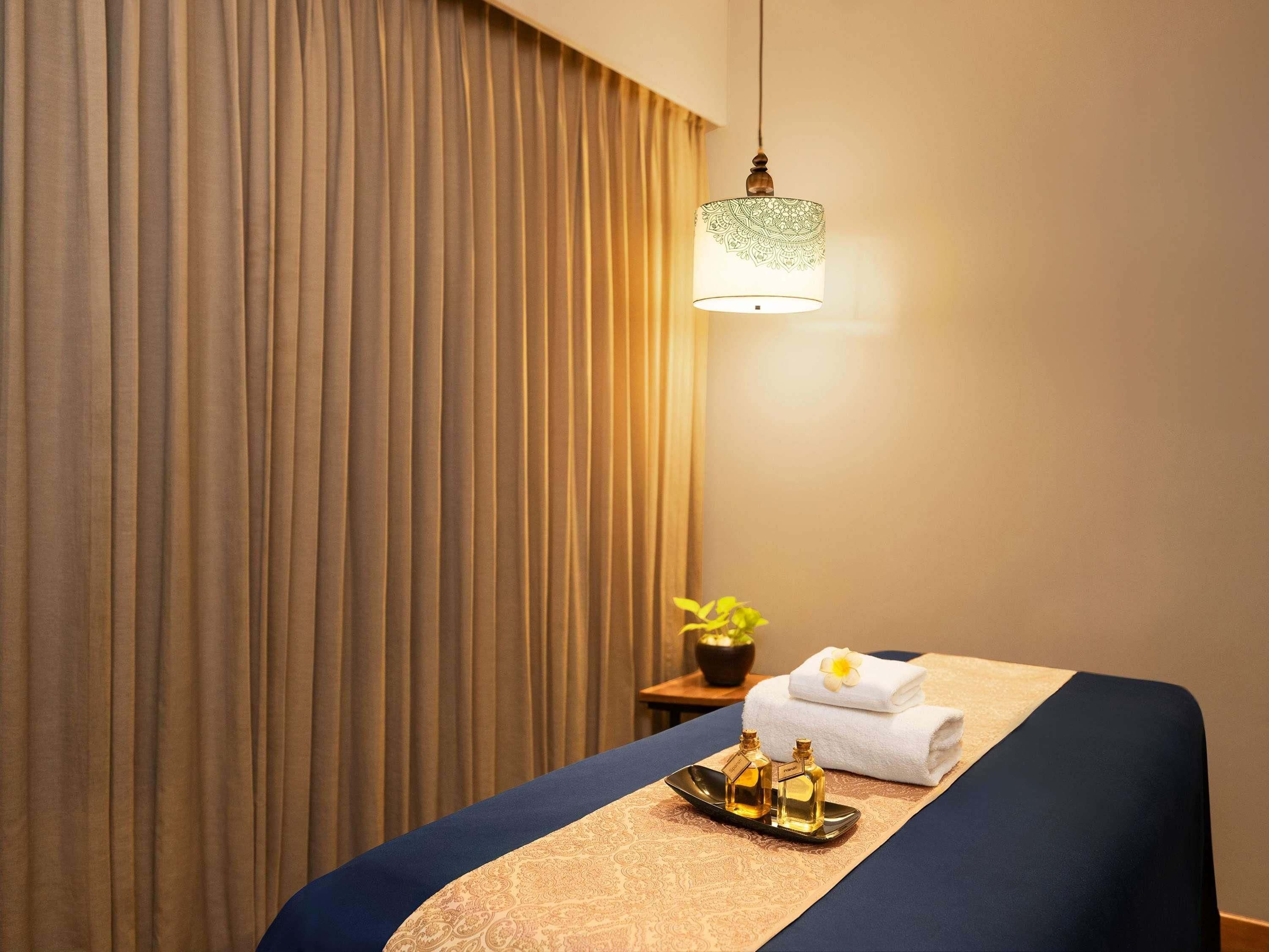 A hotel room with a bed, a bedside table with a lamp, a tray with bottles, and a towel on the bed.