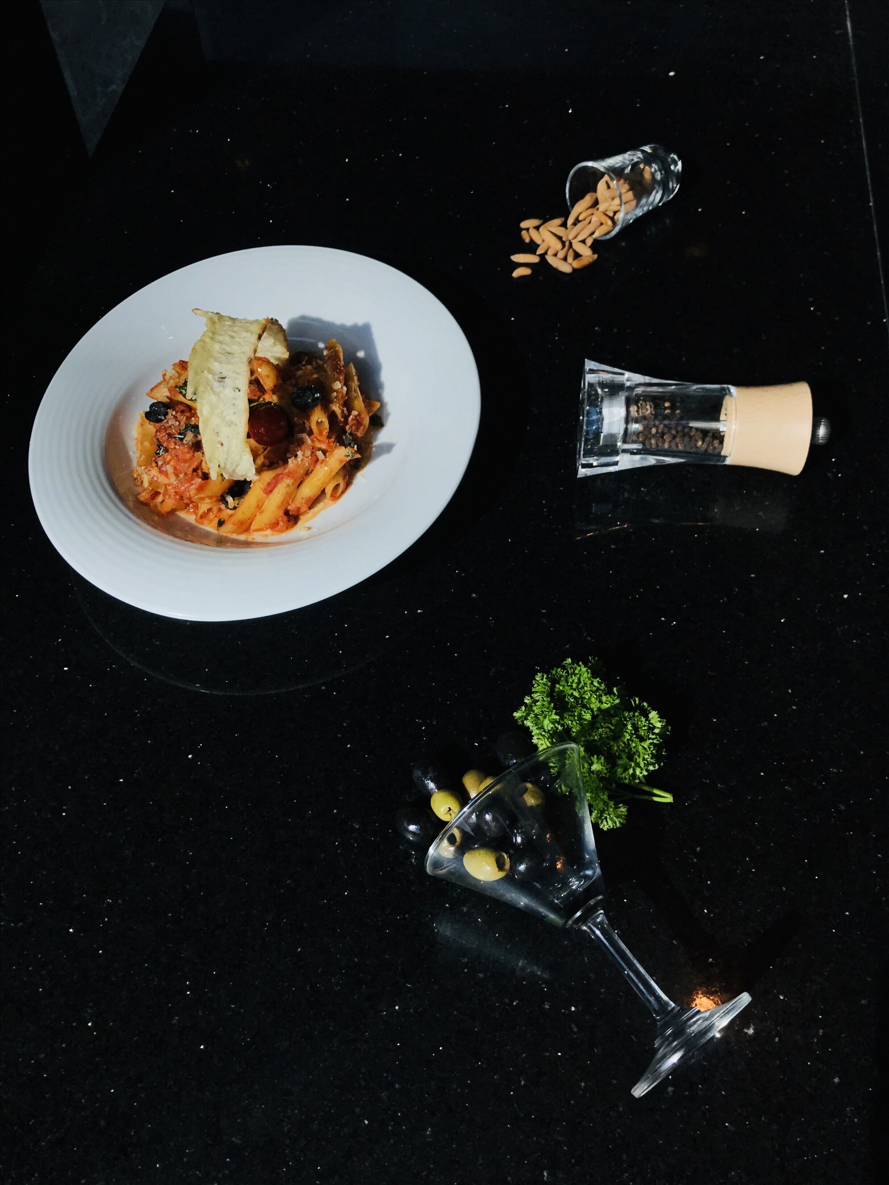 A plate of pasta with a garnish, a salt shaker, a pepper shaker, and a glass with a garnish on a dark surface.