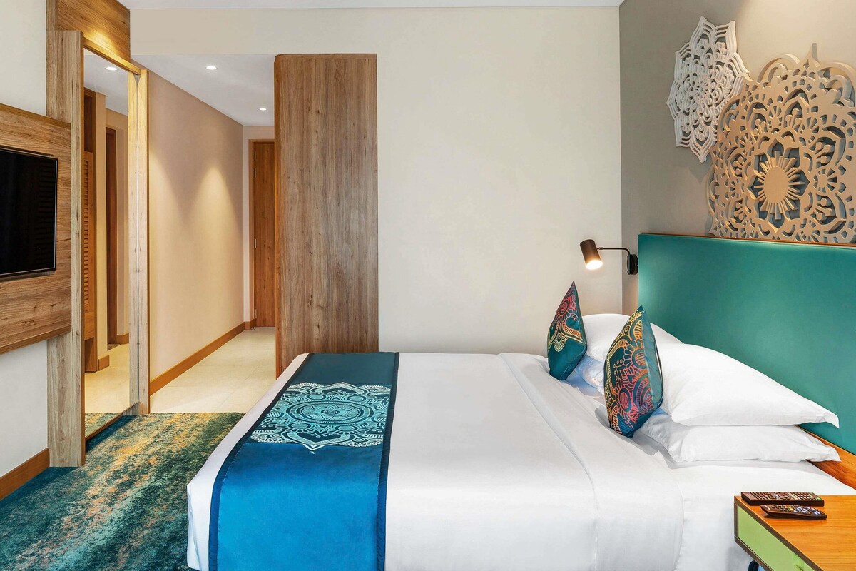 A hotel room with a bed, a TV mounted on the wall, a wooden door, and decorative wall panels.