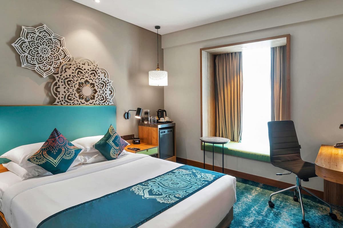 A hotel room with a bed, a desk, a chair, a window with curtains, a coffee machine, and decorative wall art.