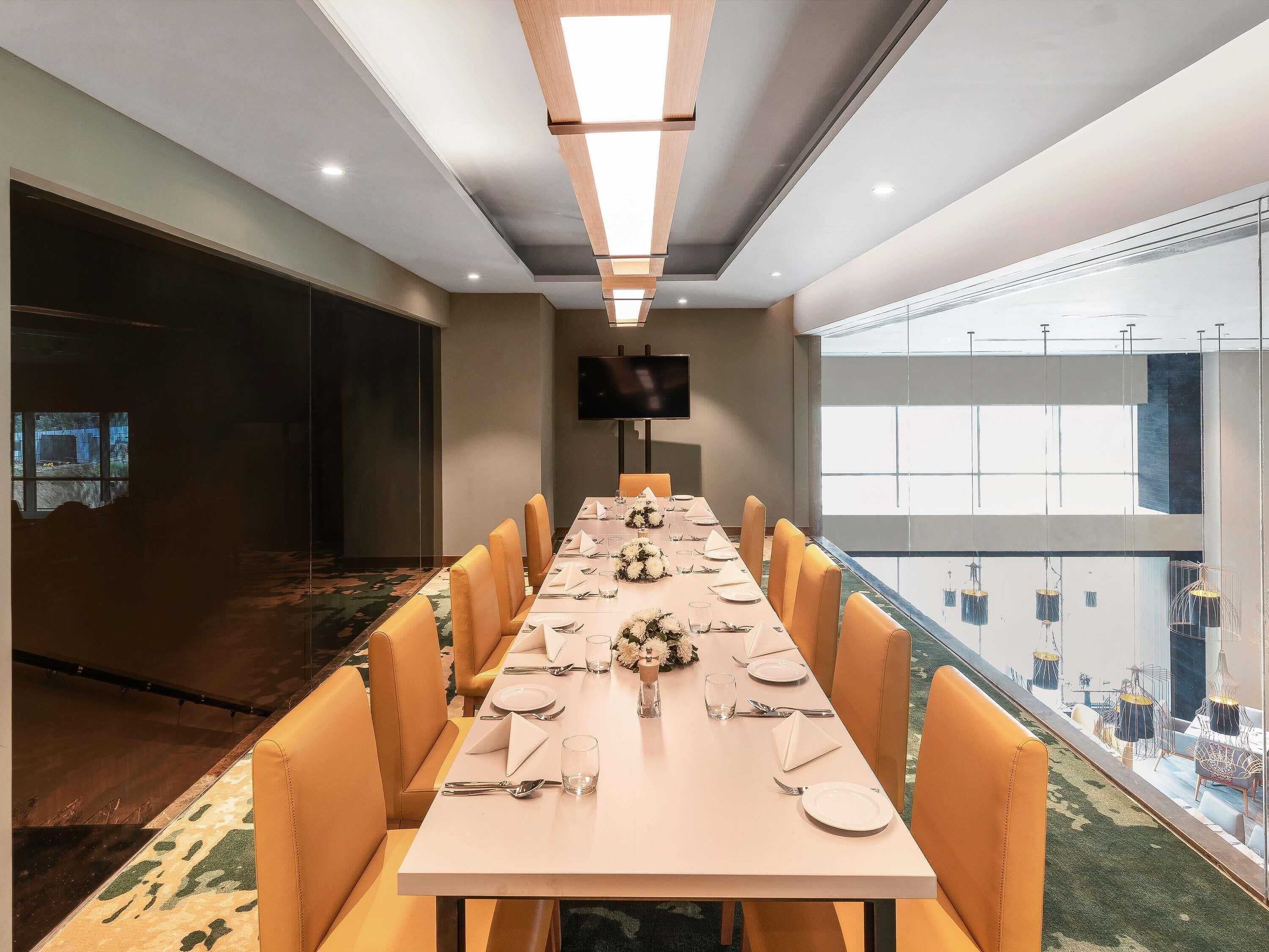 A conference room with a long table set for a meeting, equipped with chairs, a flat-screen TV, and a large window offering a view of the city.