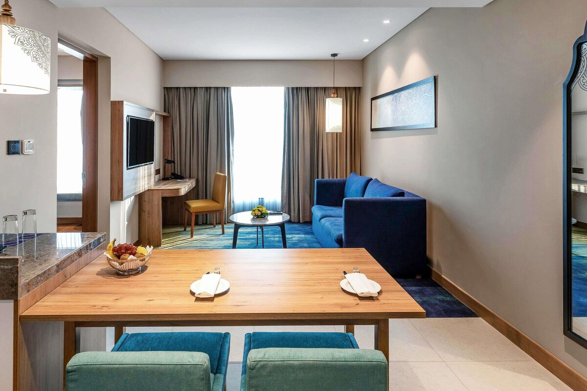 A modern hotel room with a dining area, a sofa, a TV, and a large window with curtains.