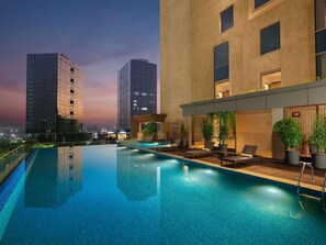 Una piscina infinita en la azotea con sillas de descanso y plantas en macetas, rodeada de edificios modernos.