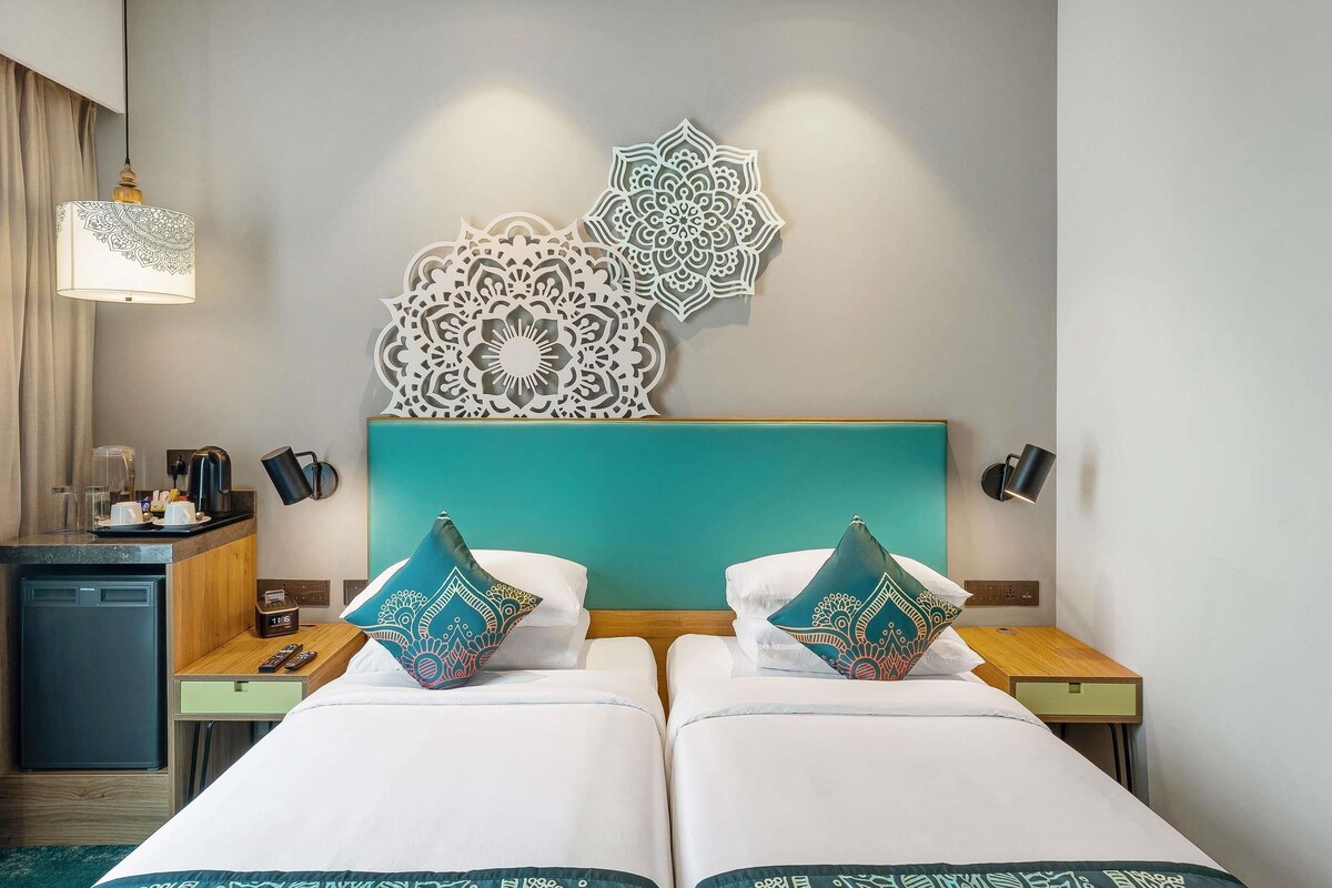 A hotel room with two beds, a small kitchenette, decorative wall art, and a bedside lamp.