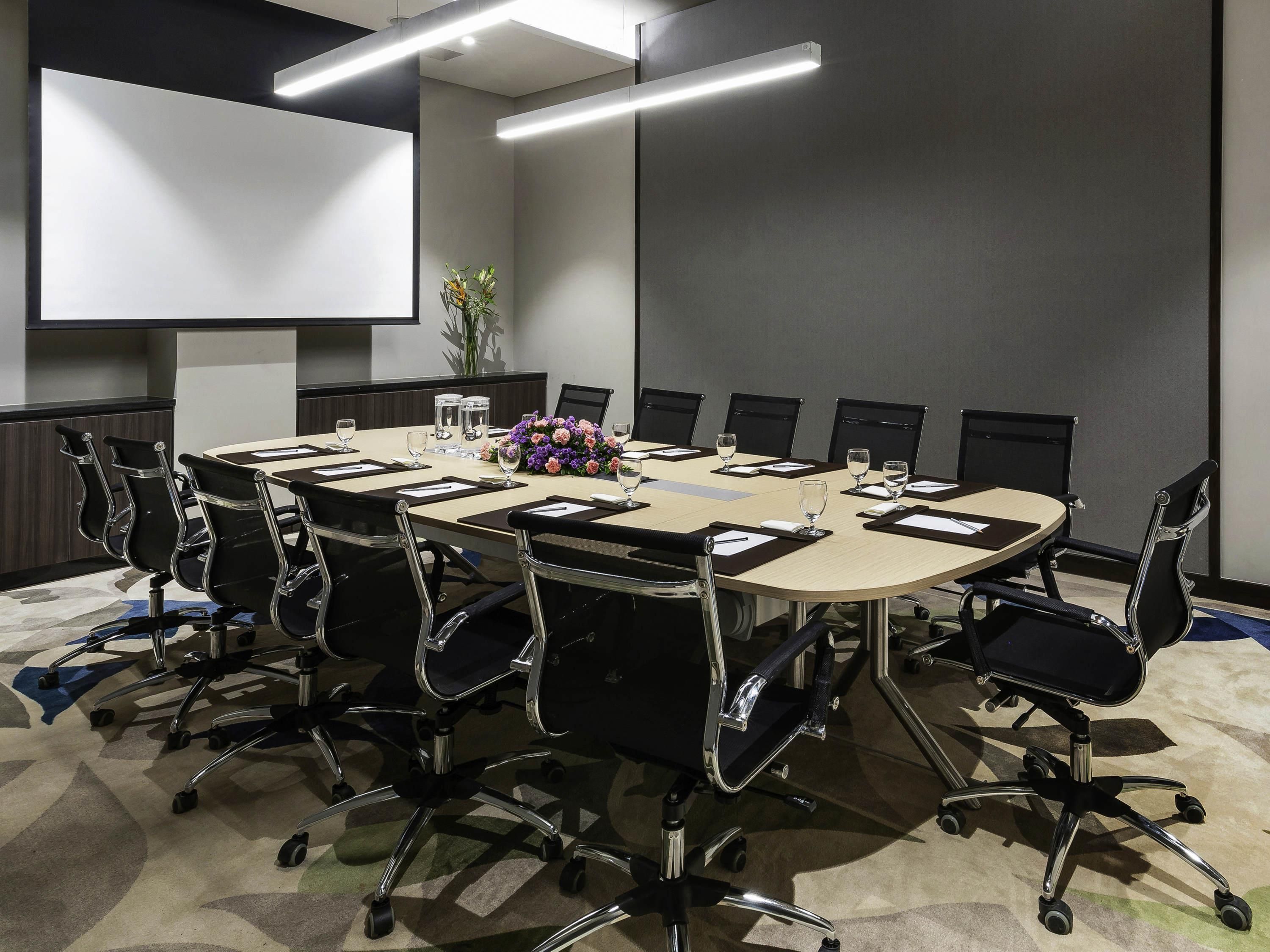 Ein Konferenzraum mit großem Tisch, Stühlen, einer Projektionsleinwand und einer Blumenkomposition.