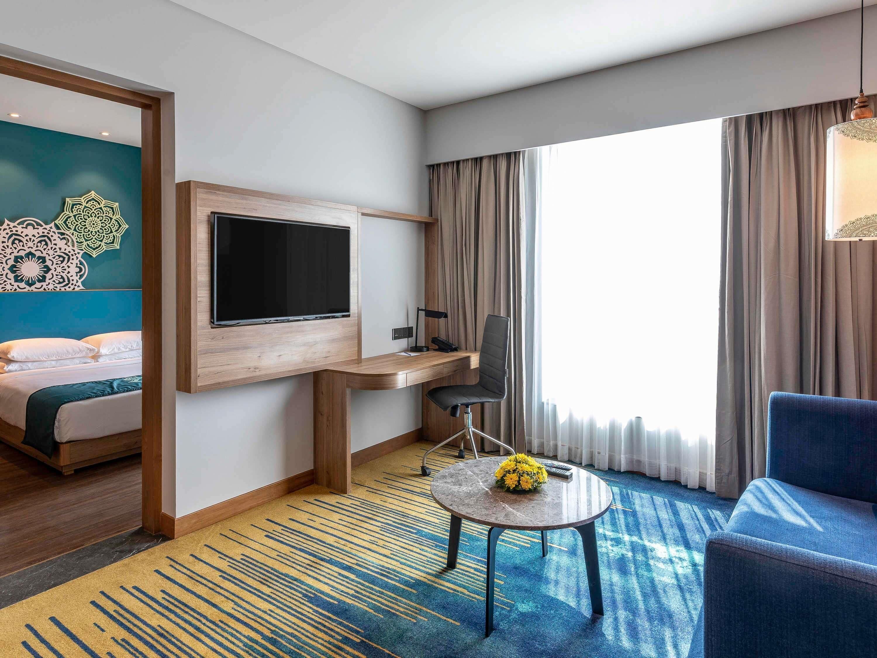 A hotel room with a bed, a desk with a computer, a TV, a chair, a small table with flowers, and a large window with curtains.