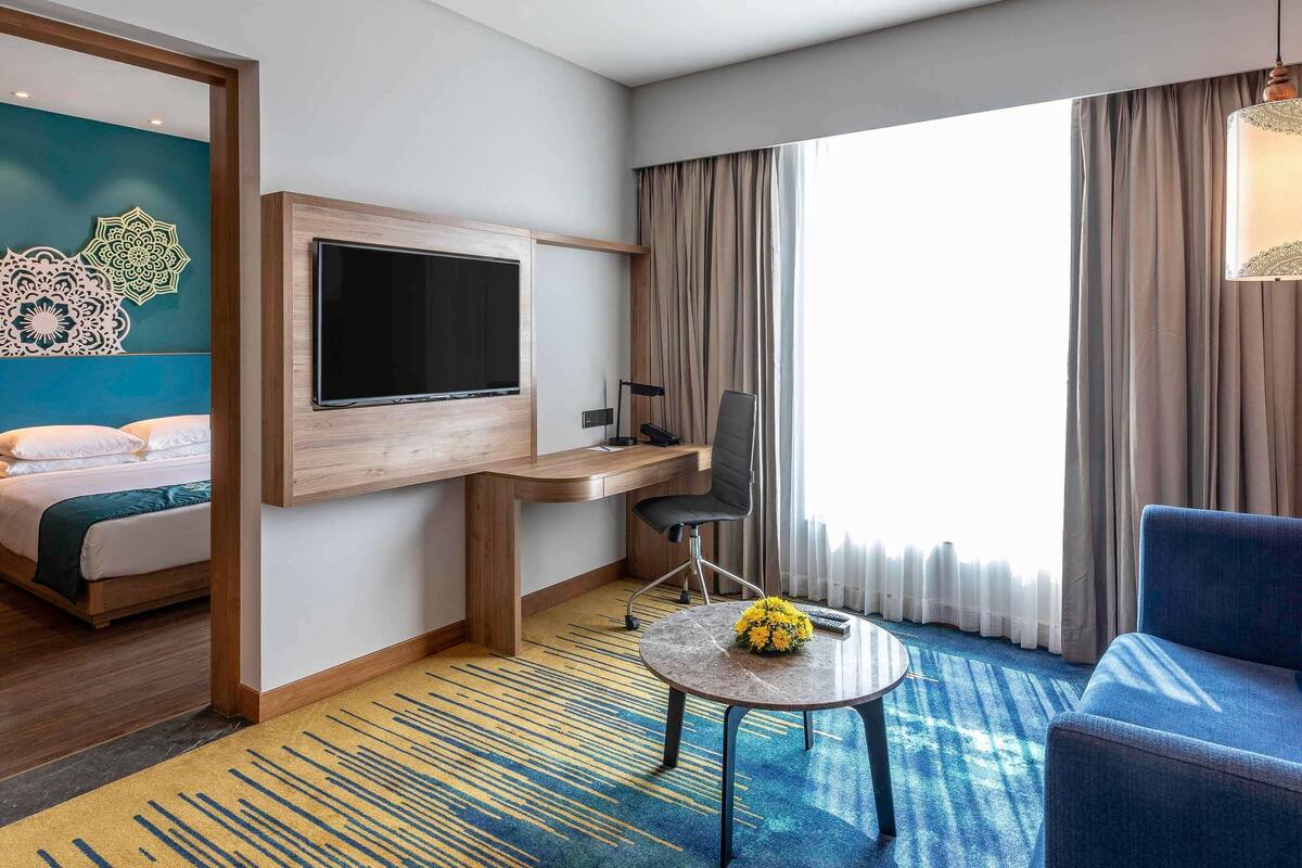 A hotel room with a bed, a desk with a computer, a TV, a chair, a small table with flowers, and a large window with curtains.