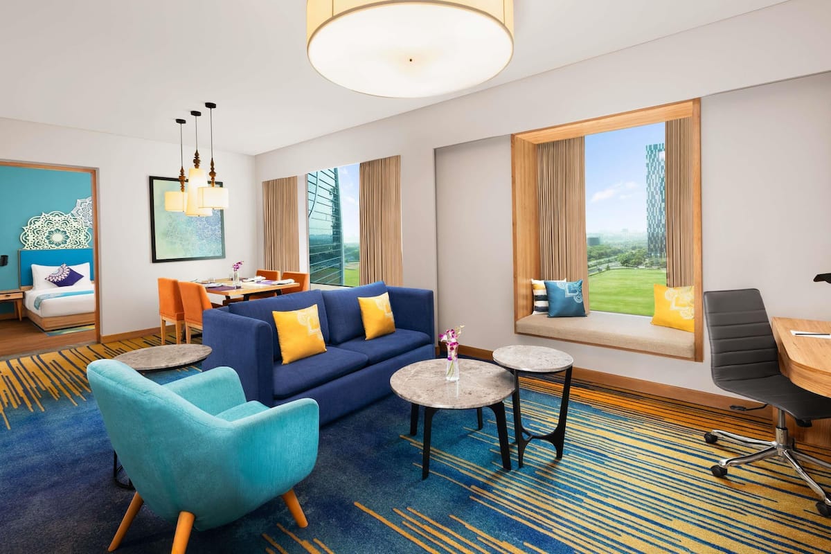 A modern hotel room with a blue sofa, teal armchair, and a dining area with a table and chairs. Large windows offer a view of the outdoors.