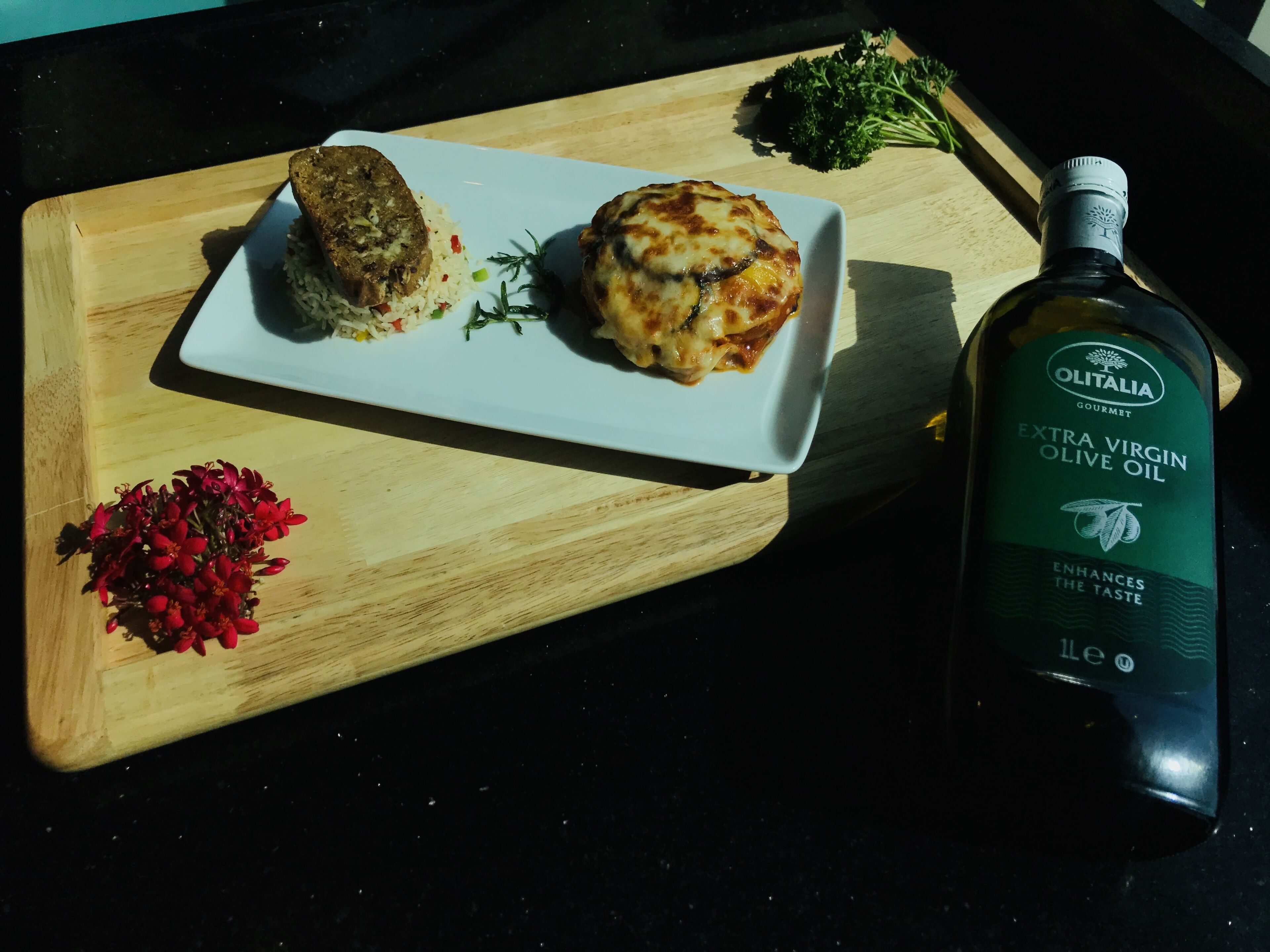 Une planche de service en bois sur laquelle se trouve une assiette de nourriture, une bouteille d’huile d’olive extra vierge et quelques herbes aromatiques.