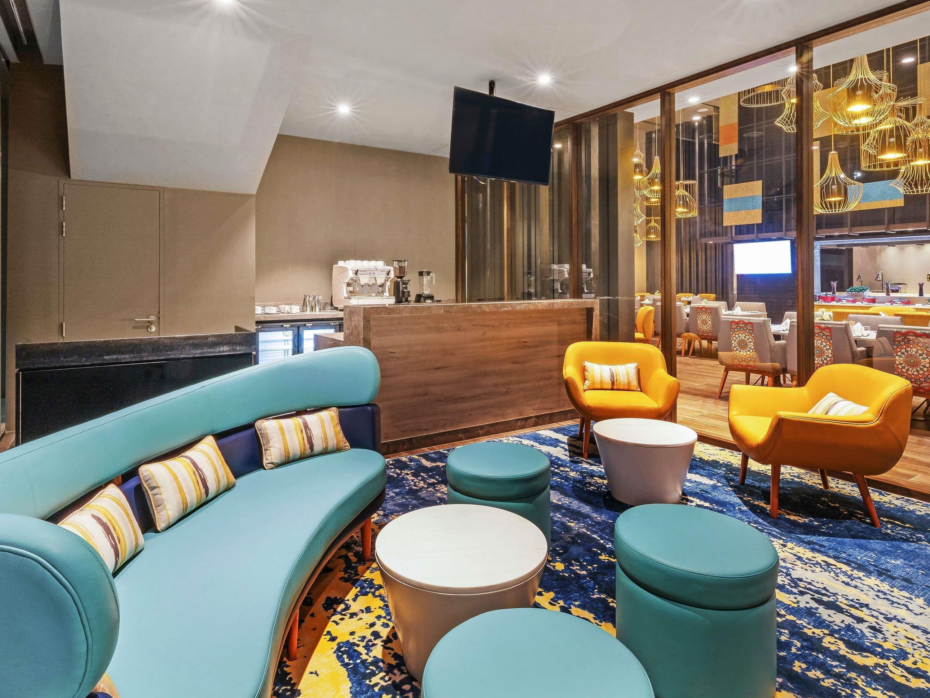 A modern lounge area with a sofa, armchairs, and ottomans, a coffee machine, and a dining area with tables and chairs.