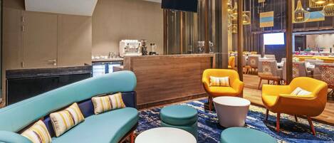 A modern lounge area with a sofa, armchairs, and ottomans, a coffee machine, and a dining area with tables and chairs.