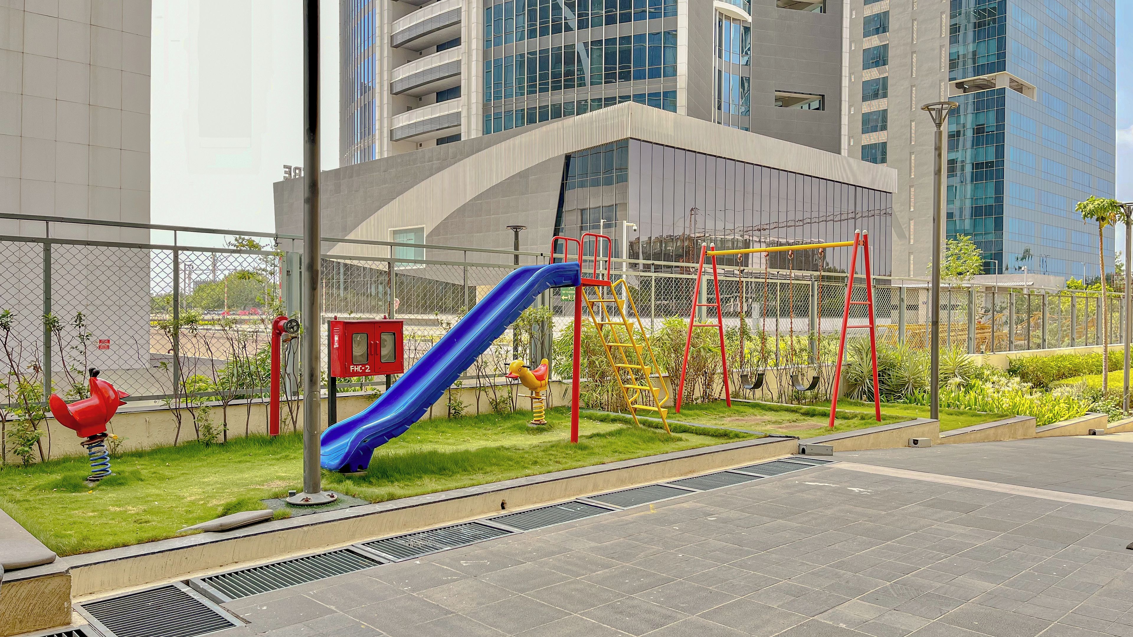 Ein Spielplatz mit einer blauen Rutsche und roten Klettergerüsten, umgeben von einem Zaun und Grünpflanzen.