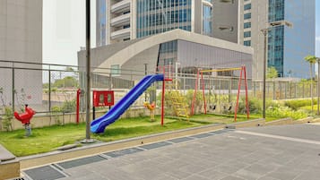 Ein Spielplatz mit einer blauen Rutsche und roten Klettergerüsten, umgeben von einem Zaun und Grünpflanzen.