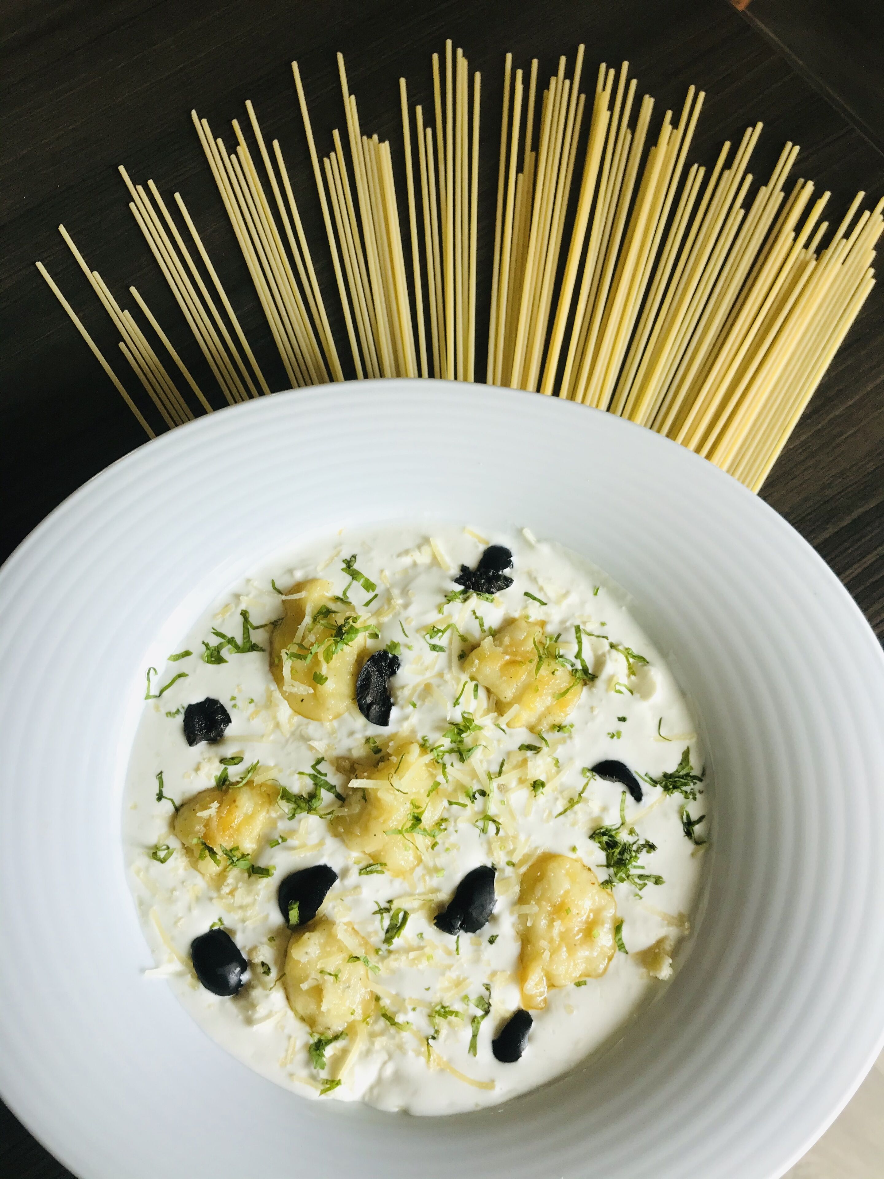 A white plate with a creamy dish garnished with herbs and olives, surrounded by uncooked spaghetti.