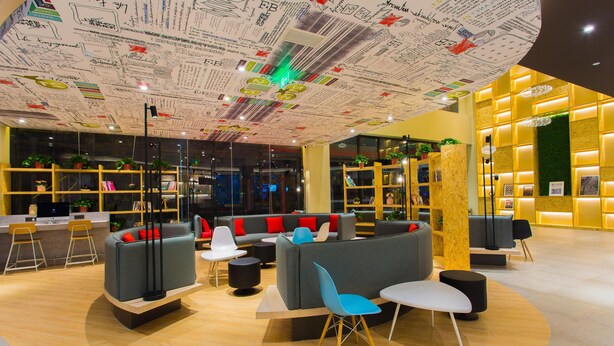 Un moderno espacio lounge con techo cubierto de inscripciones, una zona de estar con sofás y sillones, y un mostrador de recepción con pared amarilla y plantas.