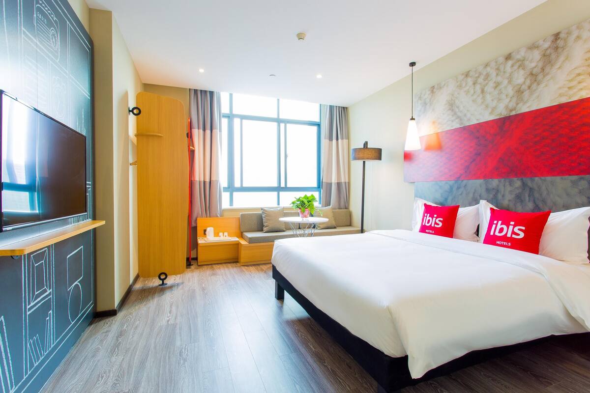 Quarto de hotel com uma cama grande, televisão, sofá, mesa pequena com planta e janela com cortinas.