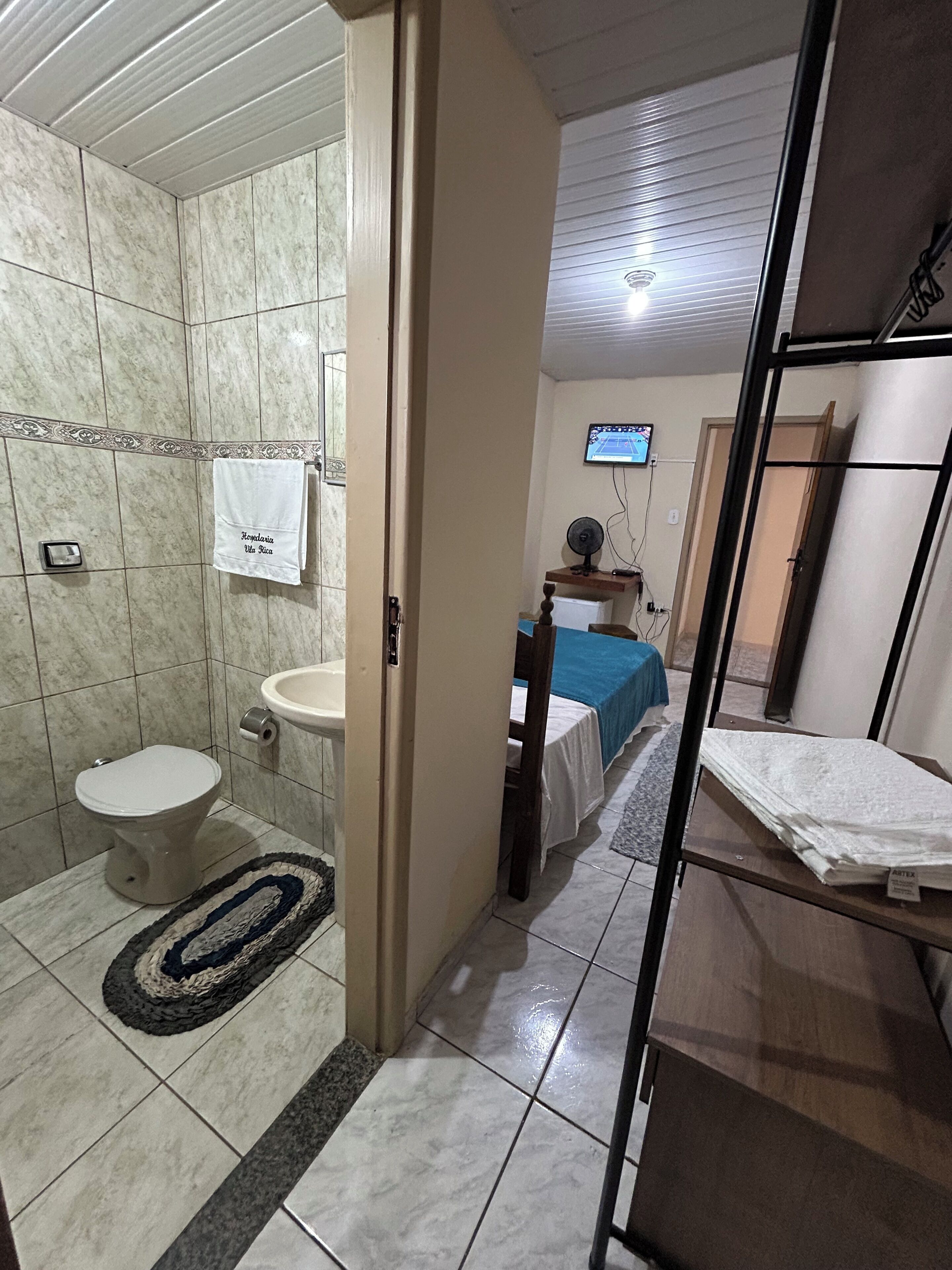 A bathroom with a toilet, sink, and shower area. There is a towel hanging on the wall and a television mounted on the wall.