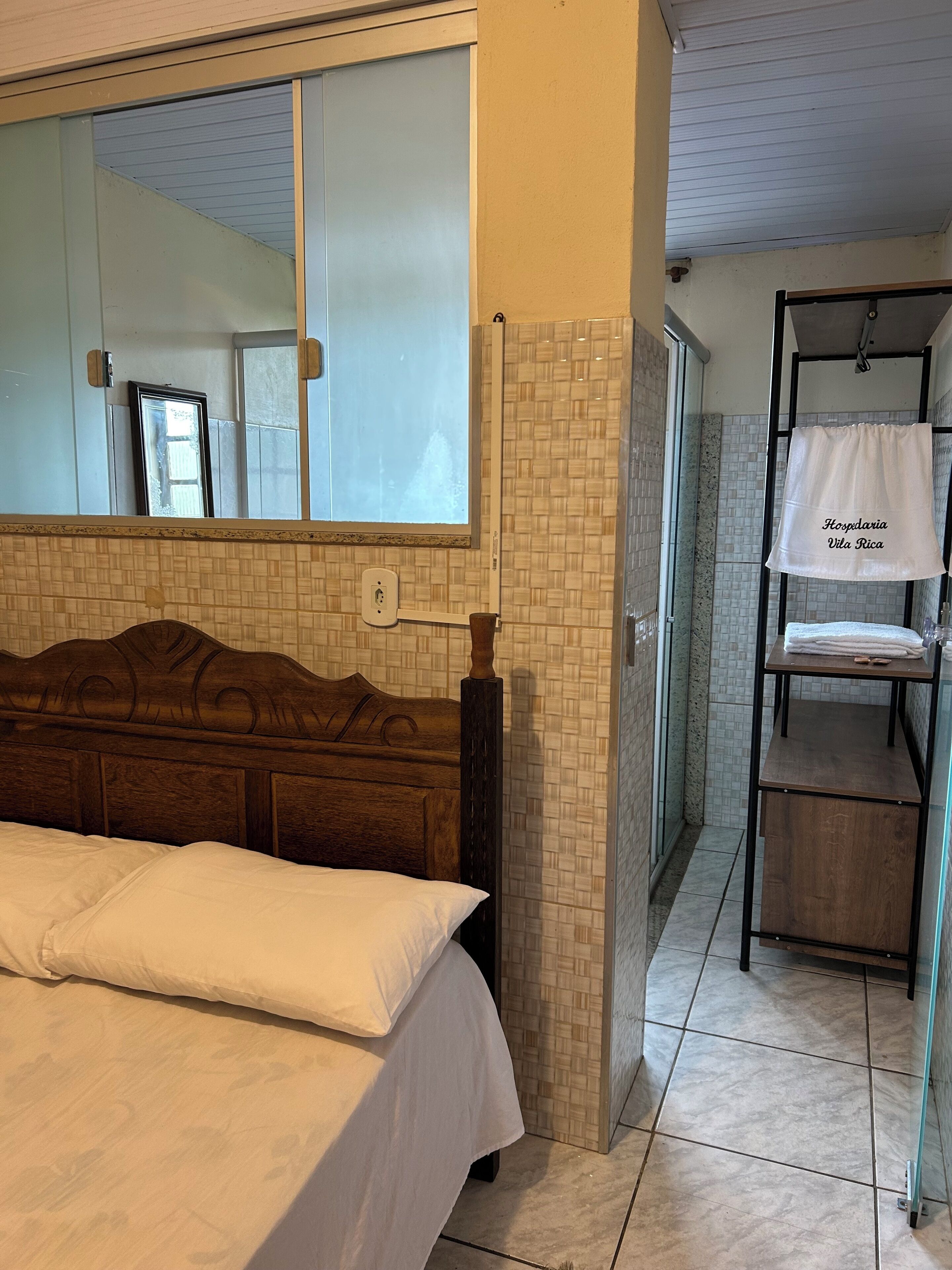 A hotel room with a bed, a wooden headboard, a bathroom with a shower, and a towel rack with a towel hanging on it.