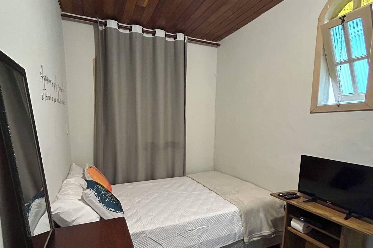 Ein kleines Schlafzimmer mit einem Bett, einem Fernseher, einer Holzkommode, einem Spiegel und einem Fenster mit Vorhängen.