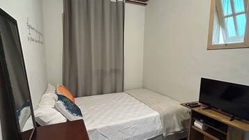 Un dormitorio pequeño con cama, televisión, cómoda de madera, espejo y ventana con cortinas.