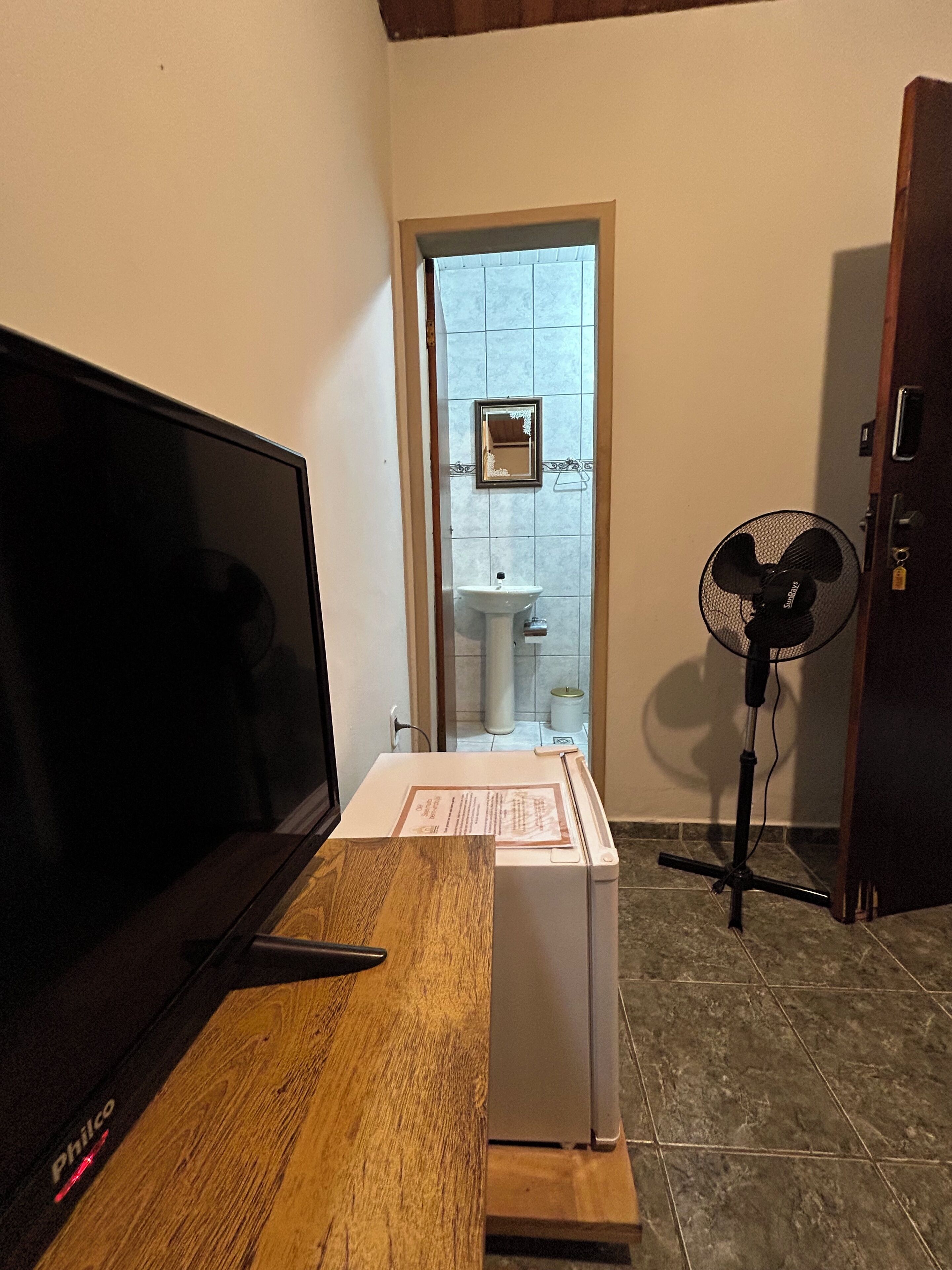 Una habitación con una televisión sobre una mesa de madera, un refrigerador blanco, un ventilador y un baño visible a través de una puerta abierta.