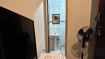 Una habitación con una televisión sobre una mesa de madera, un refrigerador blanco, un ventilador y un baño visible a través de una puerta abierta.