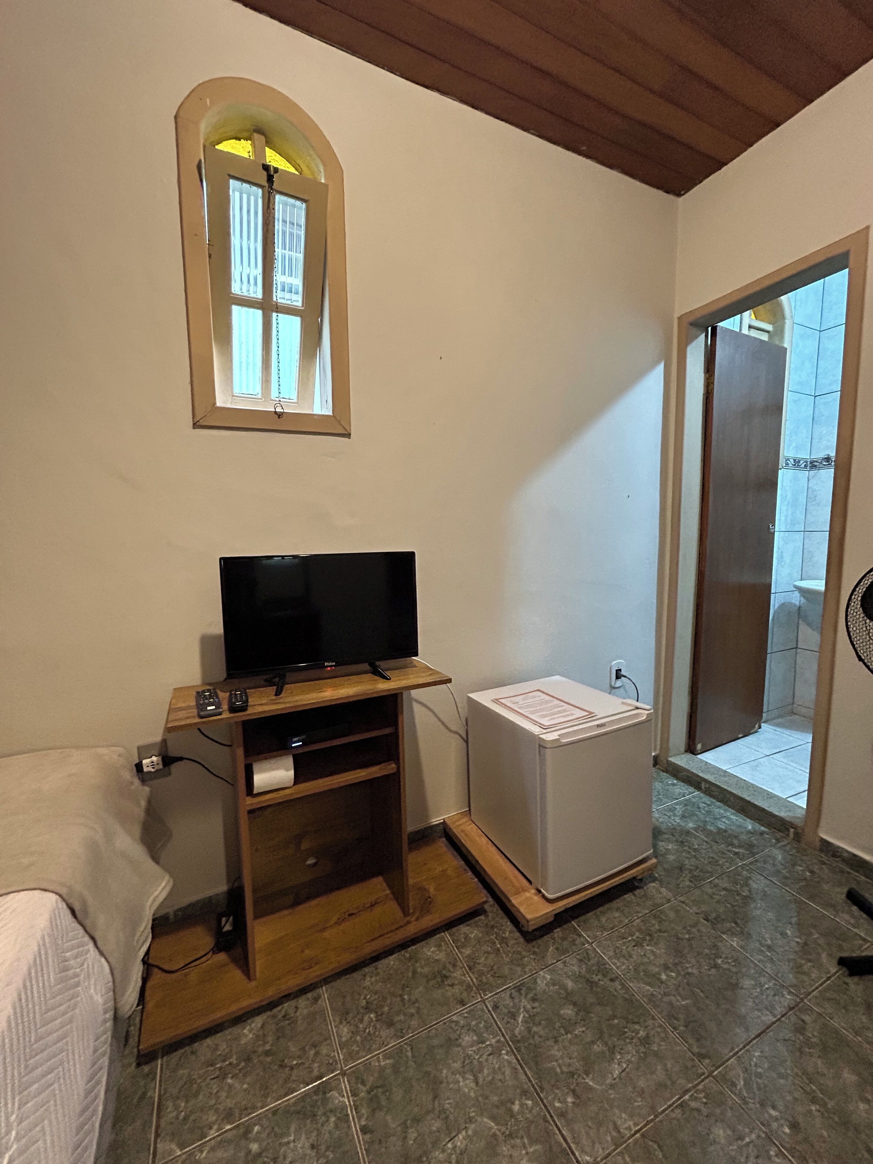 A room with a television on a wooden stand, a small white cabinet, a window with a view, and a door leading outside.