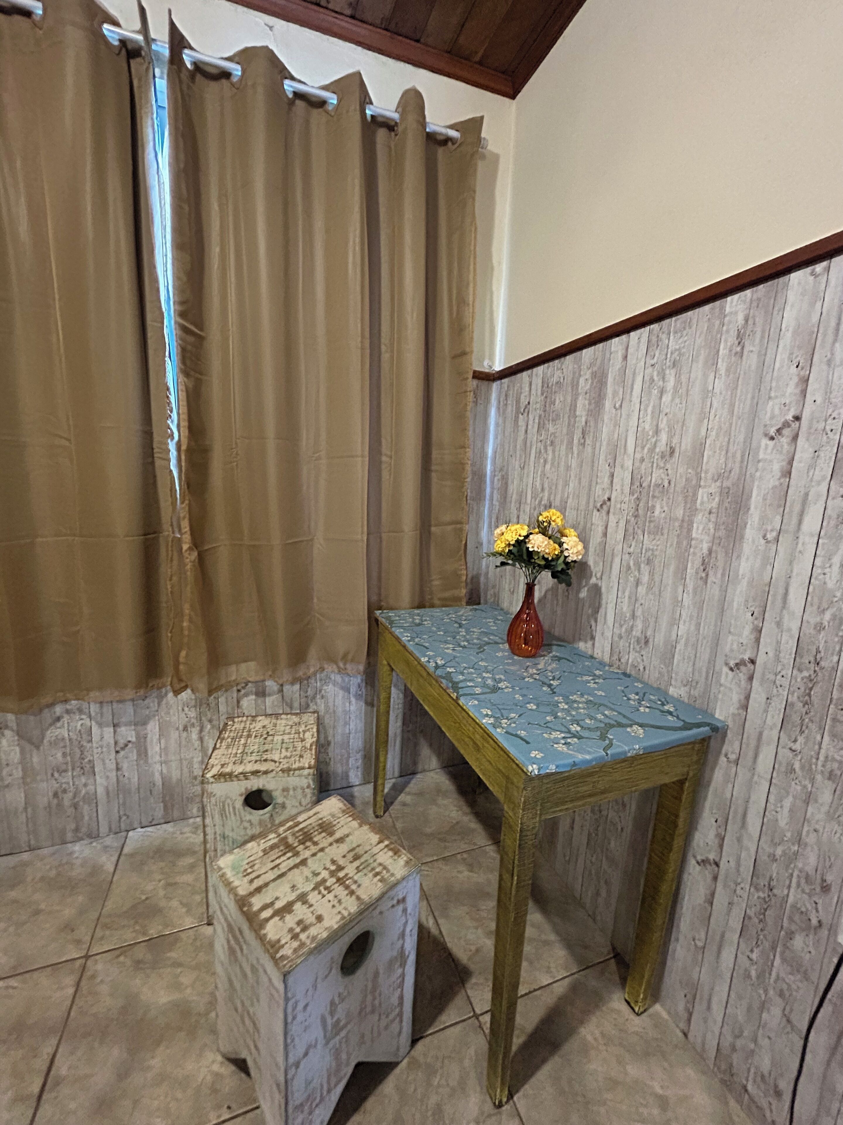 Una habitación con piso de madera, una mesita con un florero y un par de taburetes.