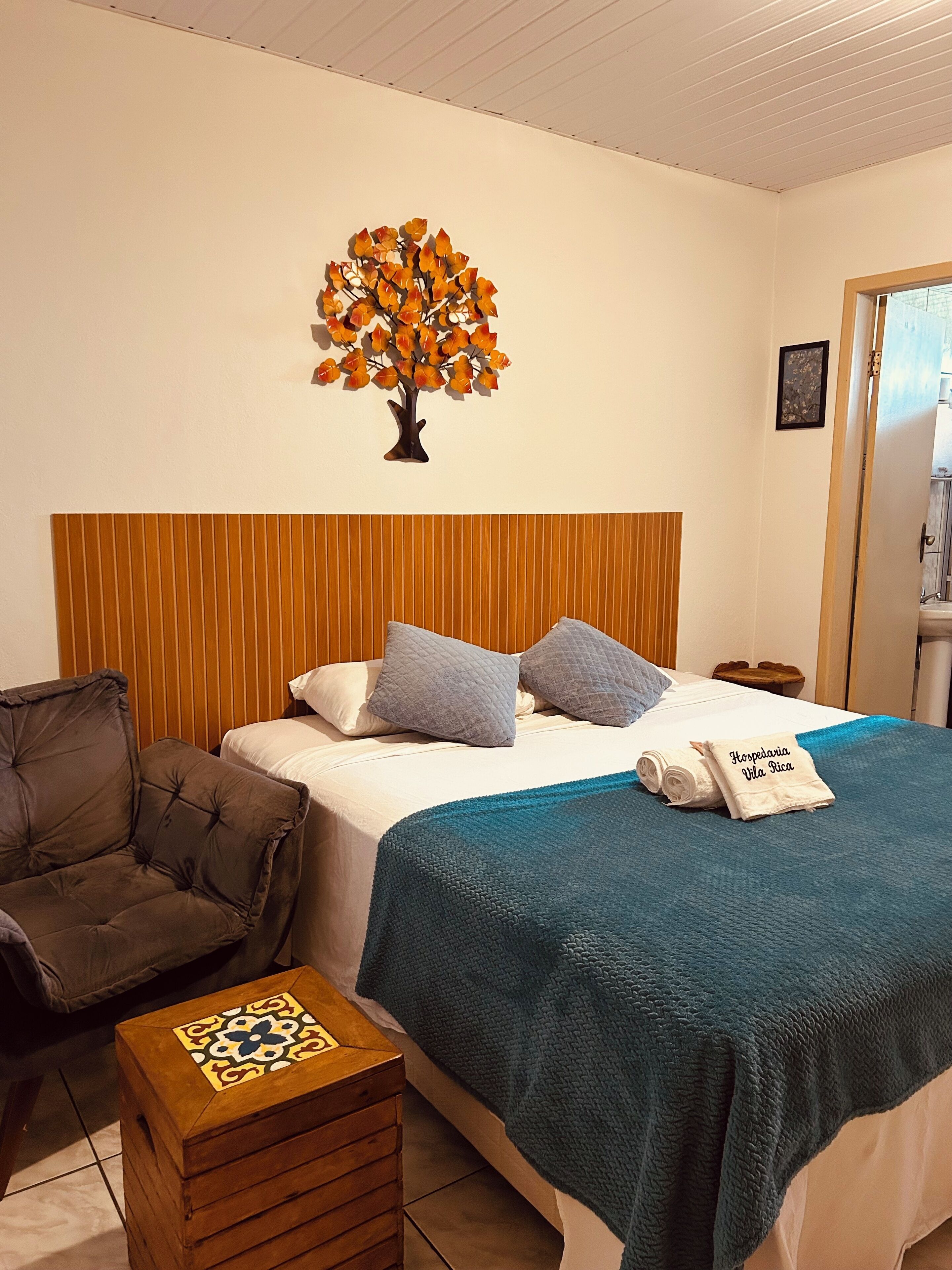 A hotel room with a bed, a wooden headboard, a decorative tree on the wall, a small wooden chest, and a chair.