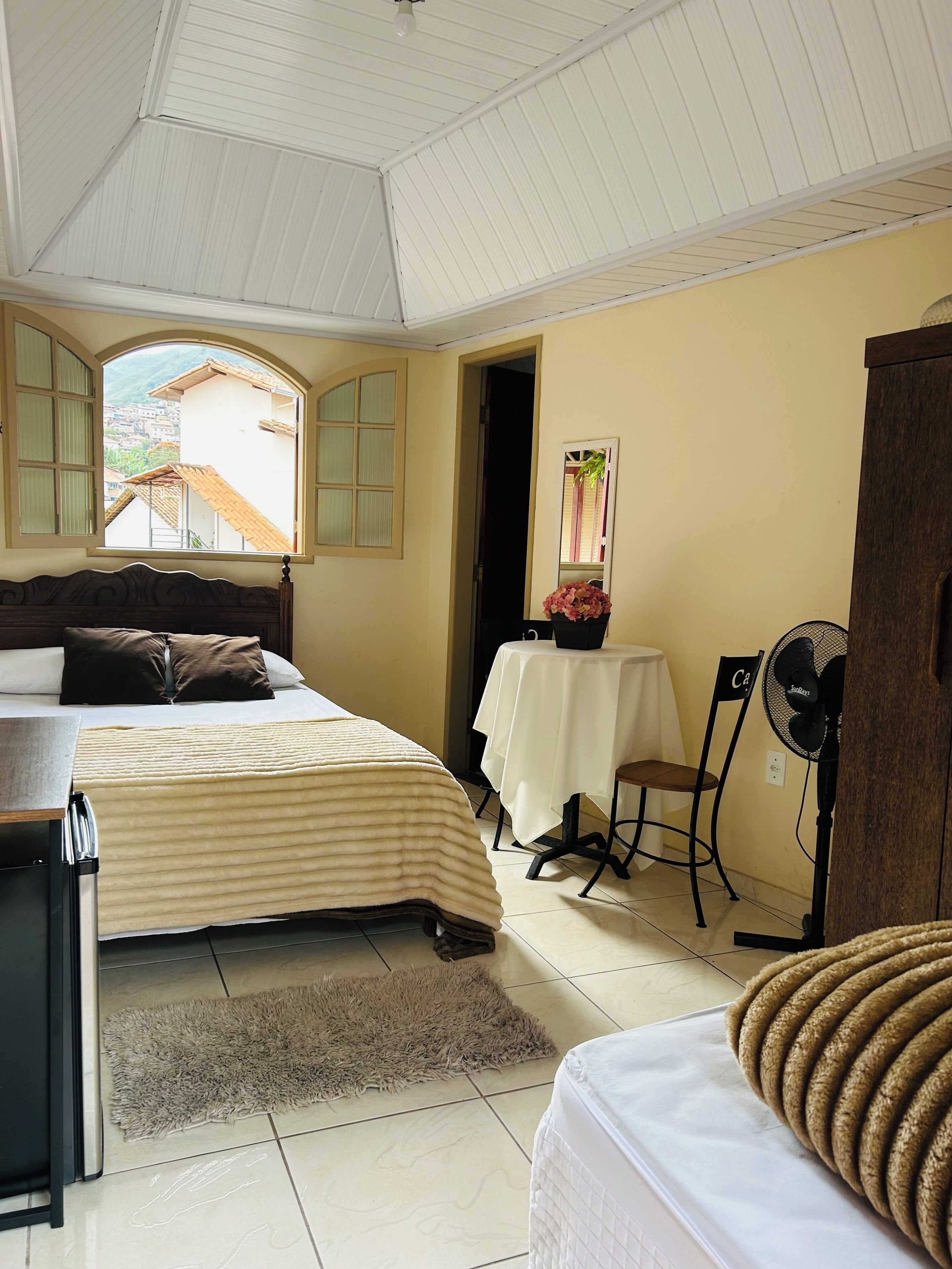 A hotel room with a bed, a desk, a chair, a table with a vase, a fan, and a view of the outside.