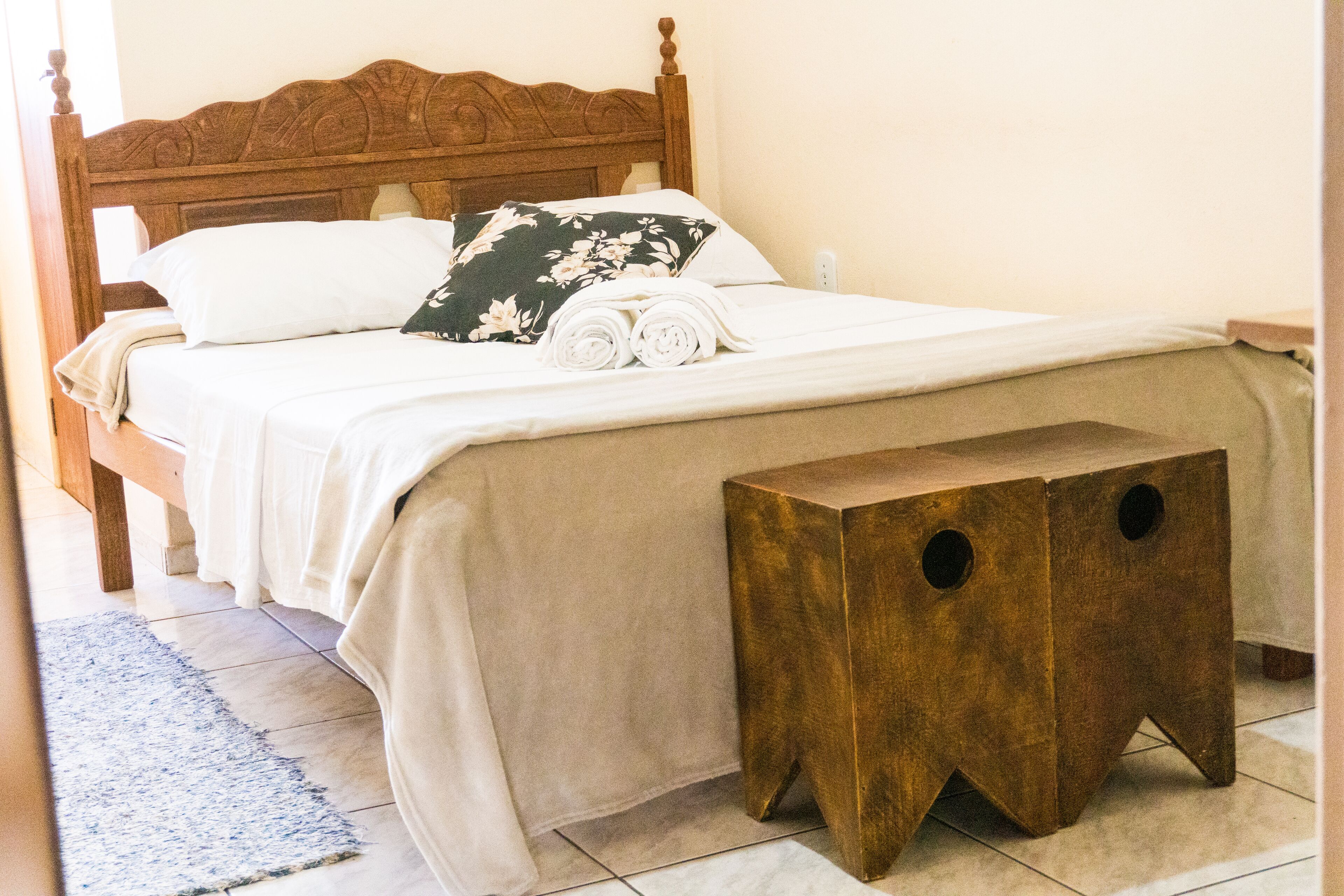 Uma cama bem arrumada com cabeceira de madeira, um pequeno banquinho de madeira e um tapete azul no chão.