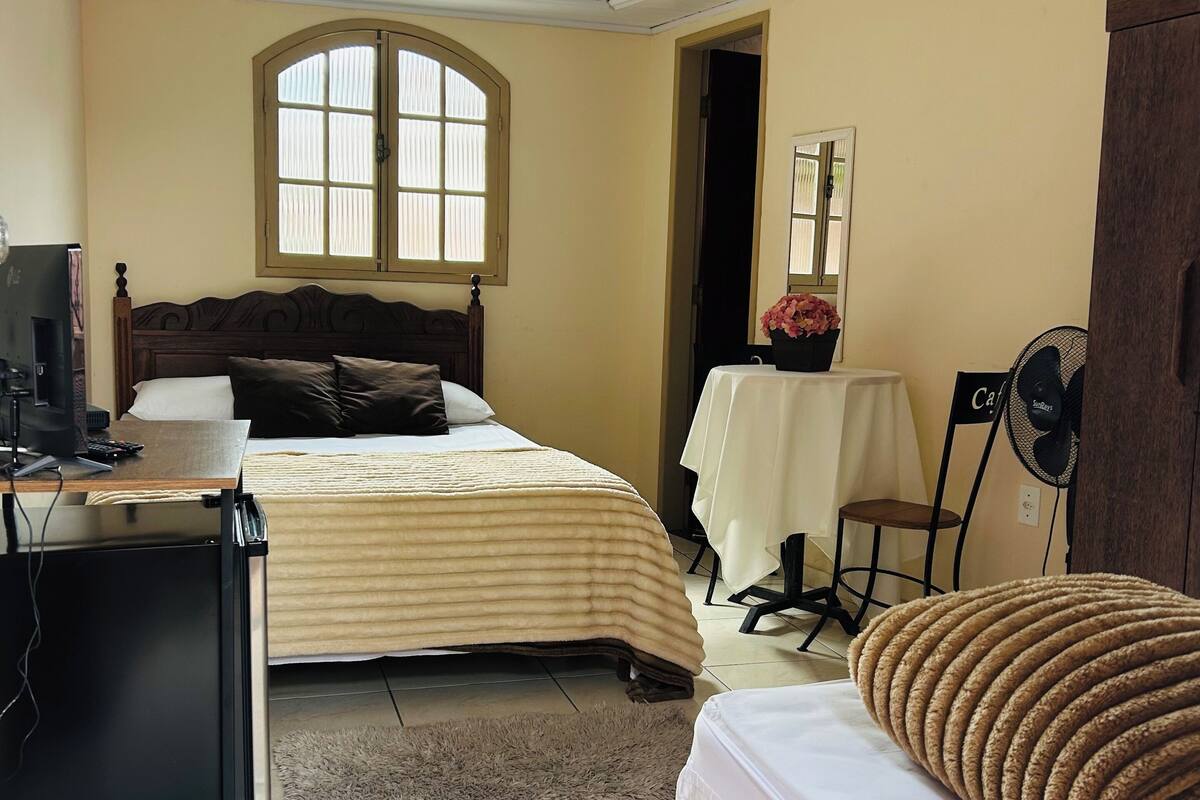 Ein Hotelzimmer mit Bett, Schreibtisch mit Computer, Stuhl, kleinem Tisch mit Ventilator und Fenster.
