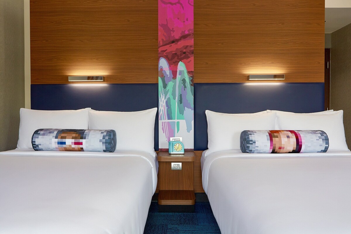 Habitación de hotel con dos camas, una mesita de noche con un reloj y un cuadro abstracto colorido en la pared.