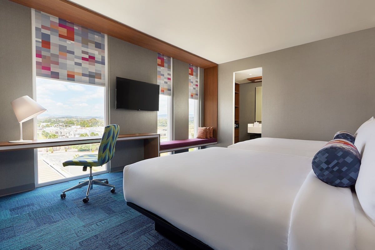 A hotel room with a large bed, a desk with a chair, a TV, and a window with a city view.