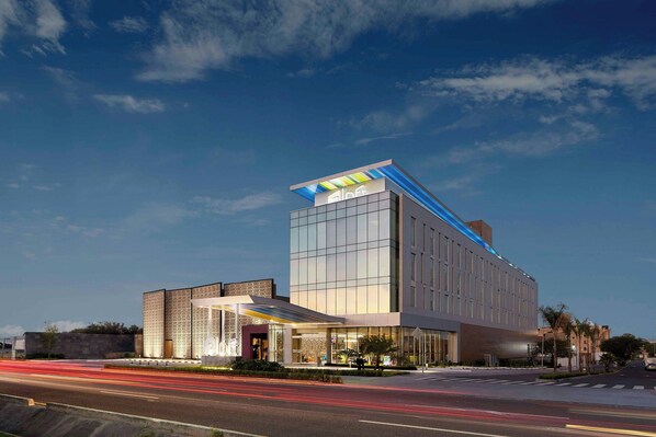 A modern hotel with a glass facade, a covered entrance, and a parking lot.