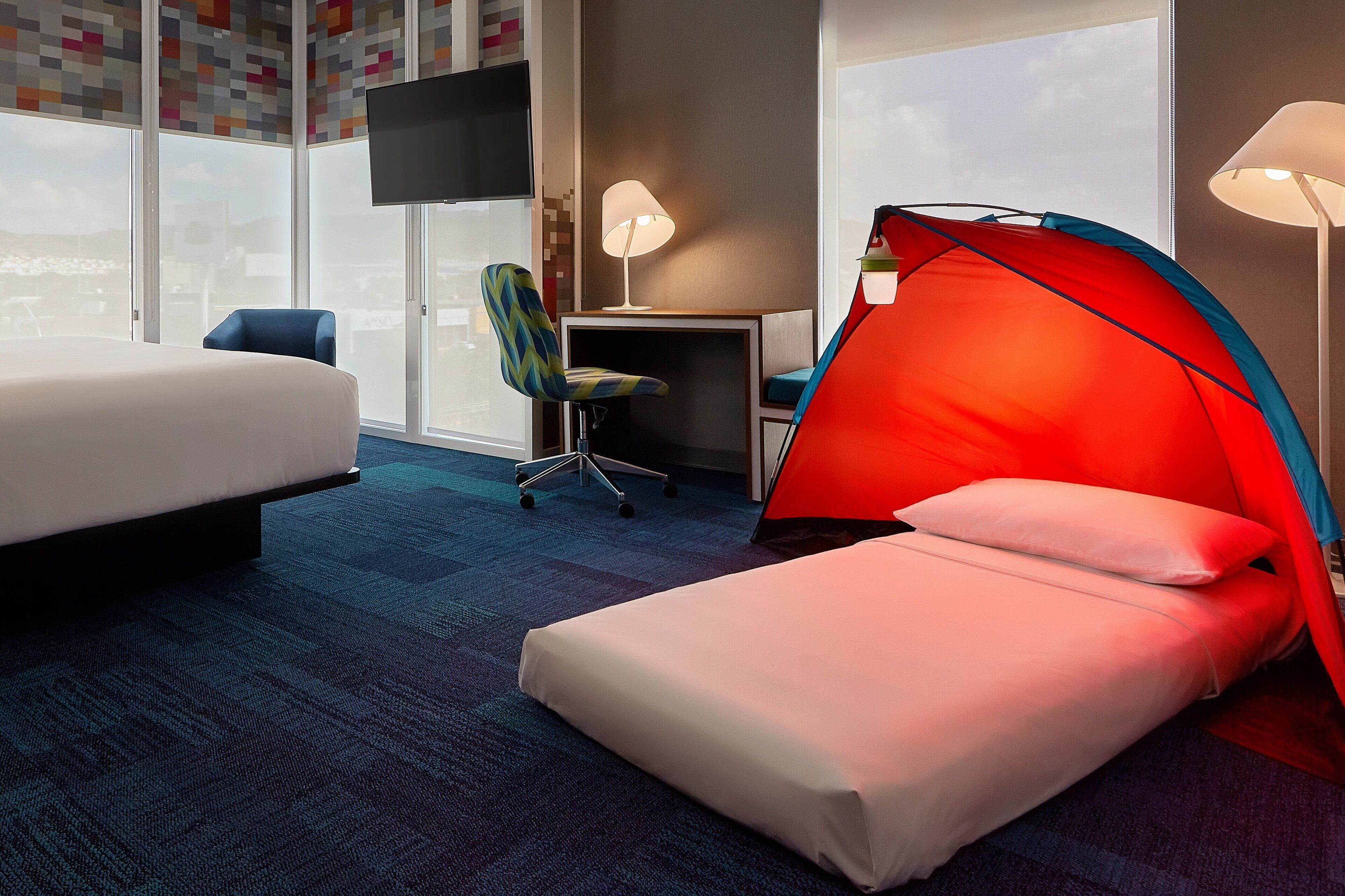 A hotel room with a bed, a desk, a chair, a tent-like structure, and a TV on the wall.