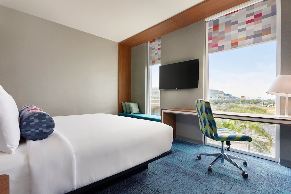 A hotel room with a bed, a desk, a chair, a TV, and a large window with a view.