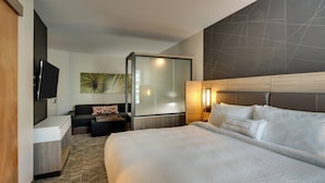 Une chambre d’hôtel avec un grand lit, un bureau doté d’un téléviseur à écran plat, un canapé et une cloison en verre.