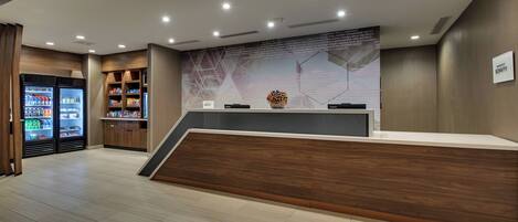 A modern reception area with a wooden reception desk, a large wall-mounted screen displaying text, and a vending machine to the left.