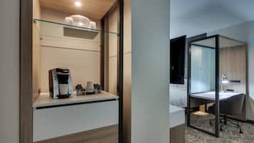 Habitación de hotel con cama, escritorio, cafetera y repisa de vidrio con un contenedor.