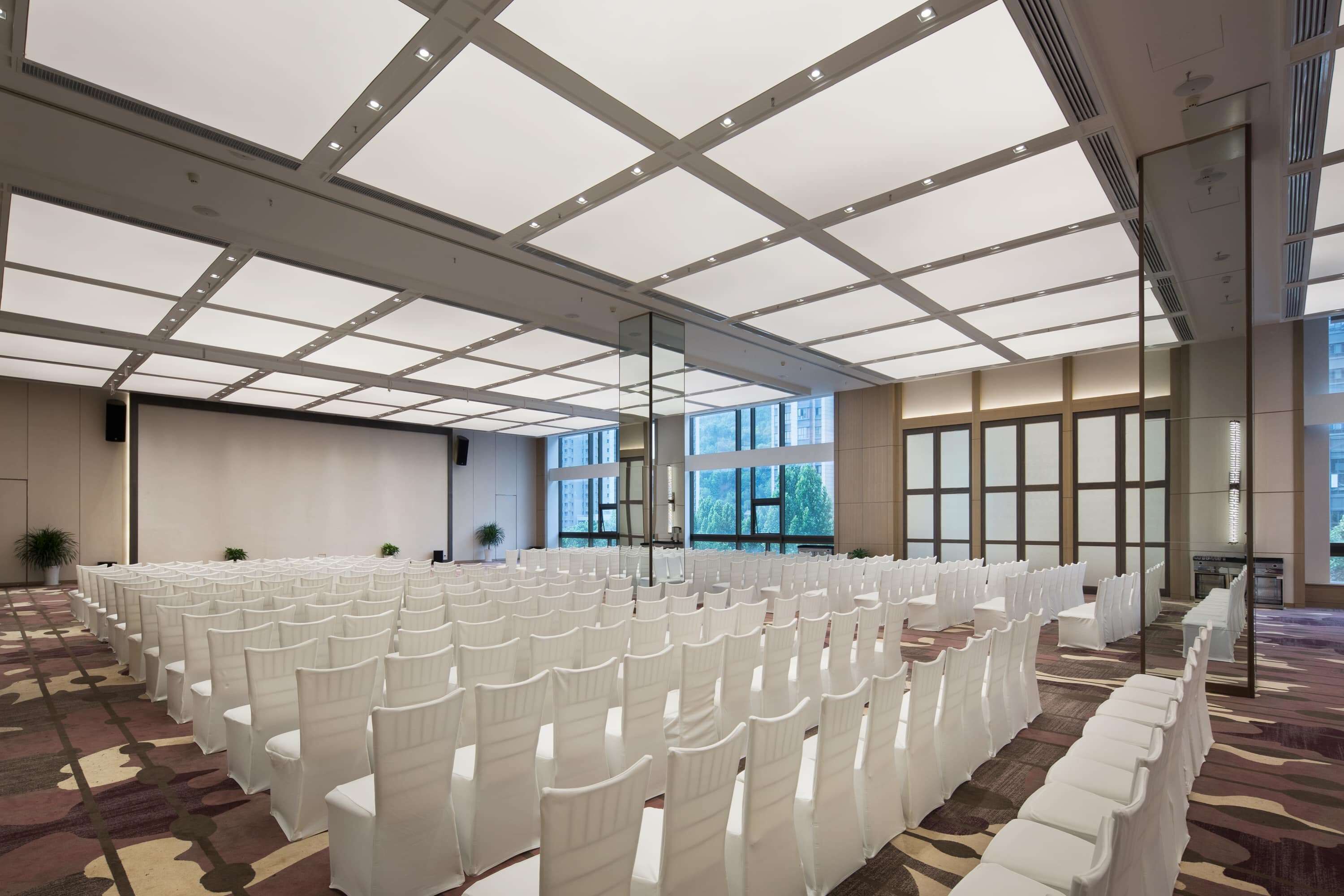 A large, well-lit room with rows of white chairs, large windows, and a patterned carpet.