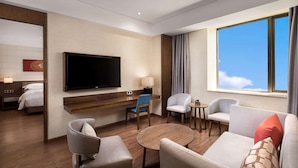 Habitación de hotel con televisión de pantalla plana, un sofá, dos sillones, una mesita de café, una mesita auxiliar pequeña y una ventana con vistas al cielo.