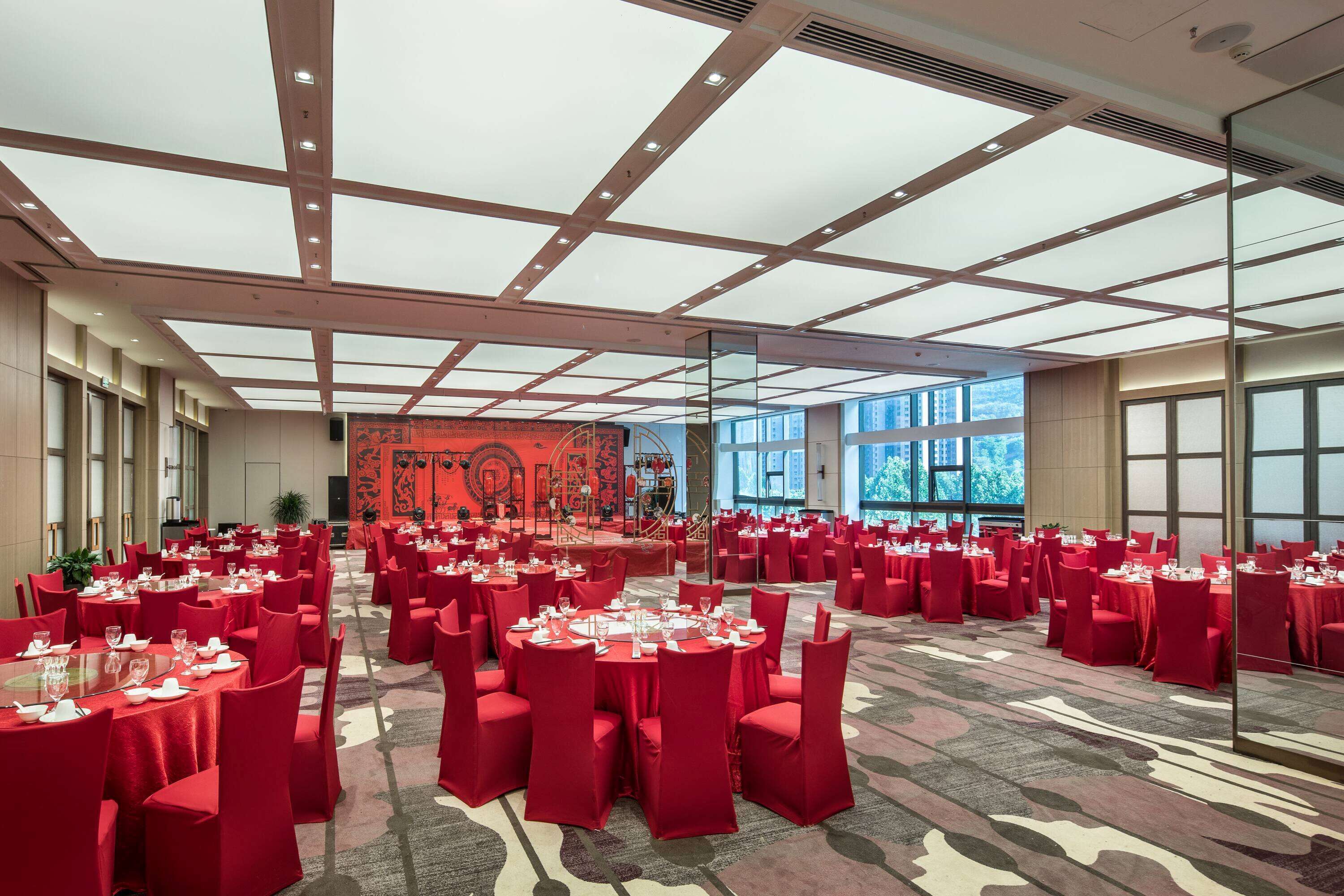 Un amplio salón de banquetes con mesas redondas y sillas rojas, un escenario con un telón rojo y grandes ventanales que ofrecen vistas de la ciudad.