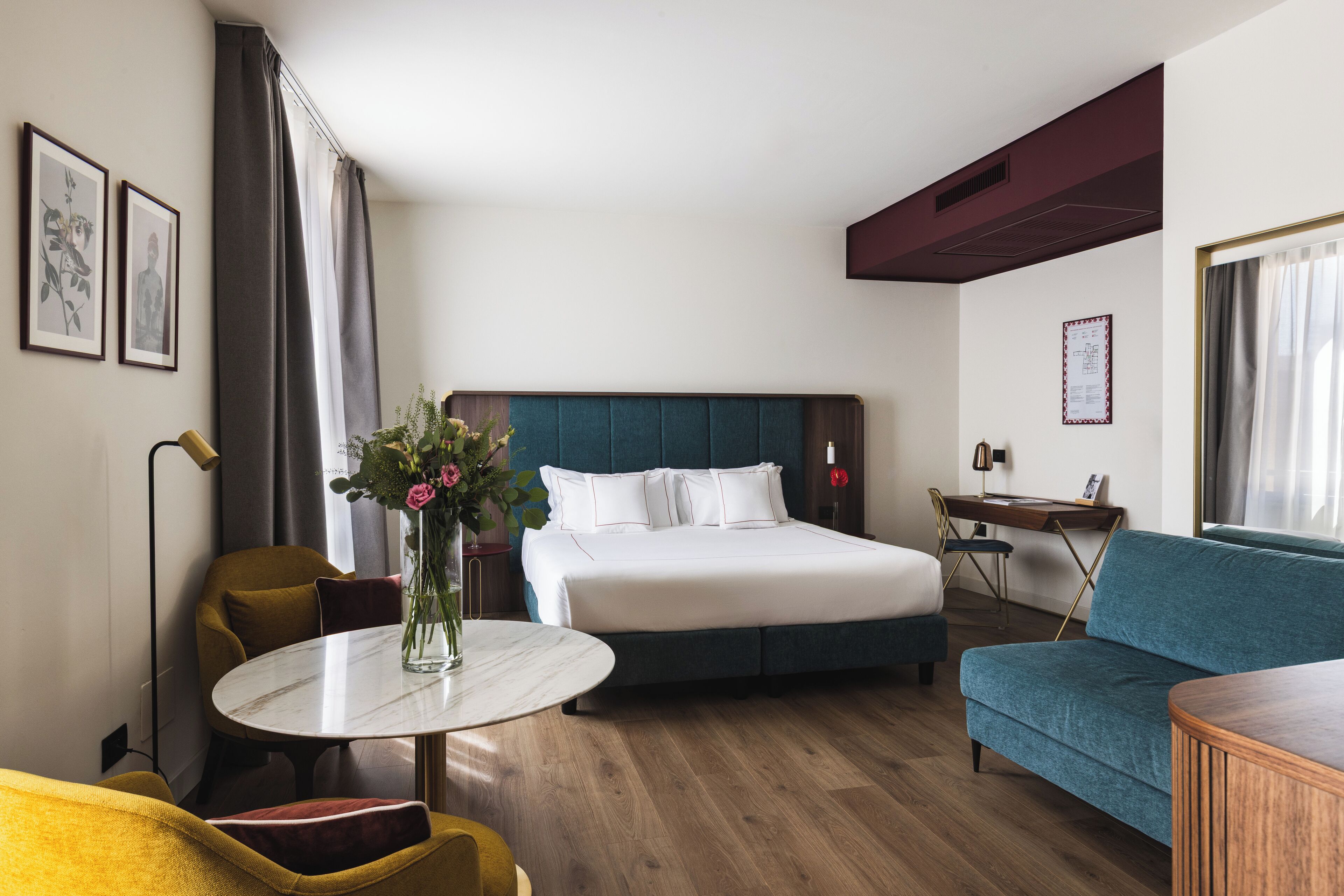 A hotel room with a bed, a sofa, a round table with a vase, a desk, and a chair.