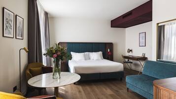 A hotel room with a bed, a sofa, a round table with a vase, a desk, and a chair.
