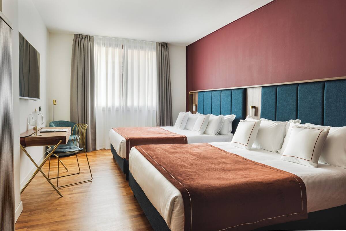 A hotel room with two beds, a desk, a chair, a TV, and a large window with curtains.